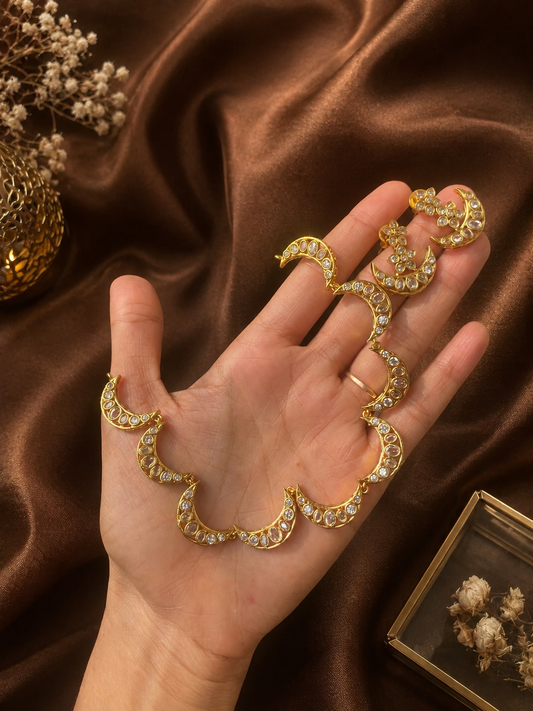 A hand displaying a gold-plated choker necklace and matching stud earrings set, styled with a continuous chain of linked crescent moon motifs embedded with clear cz stones against a rich brown satin fabric.