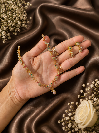 A hand holding a delicate, slender gold-plated vine-style necklace and matching drop earrings, detailed with tiny white, ruby red, and emerald green AD stones on a shimmering brown satin backdrop.