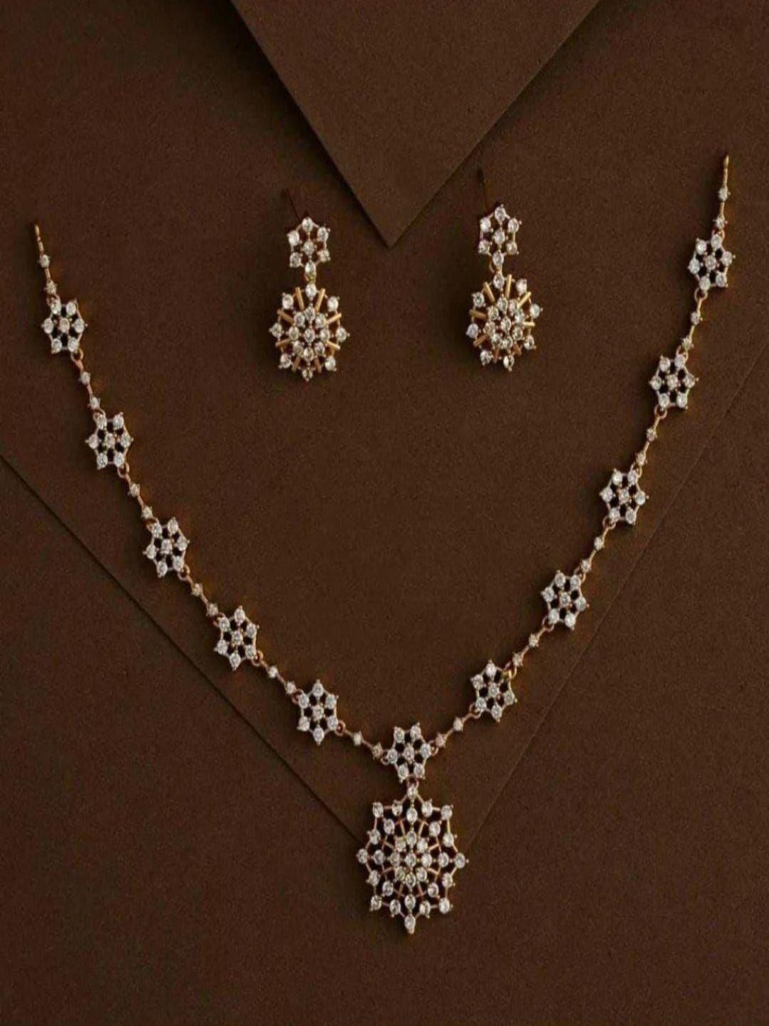 A delicate gold-plated necklace set displayed on a dark brown background, featuring a chain of small, interconnected white CZ stone clusters leading to a larger snowflake-shaped starburst pendant with matching stud earrings.