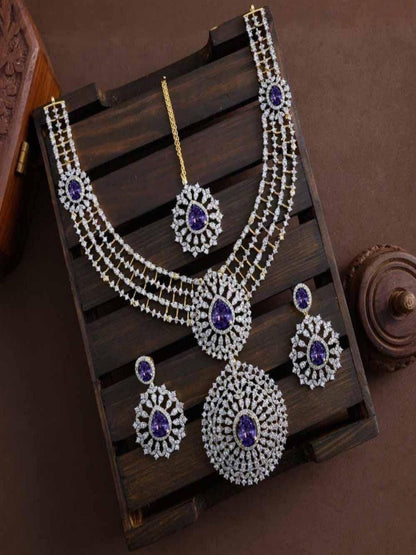 A grand bridal jewelry set displayed on a dark wooden surface, including a 4-row gold-plated white CZ necklace with a large circular ruby pink statement pendant, matching drop earrings, and a coordinating maang tikka.