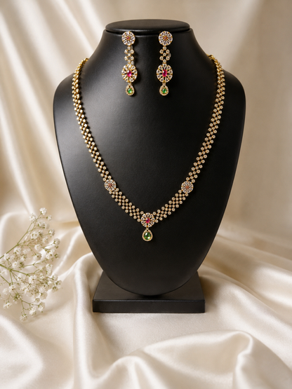 A long, double-layered gold-plated haraam necklace and matching drop earrings displayed on a black jewelry bust, featuring delicate floral motifs set with white, ruby red, and teardrop emerald green AD stones against a cream satin backdrop.