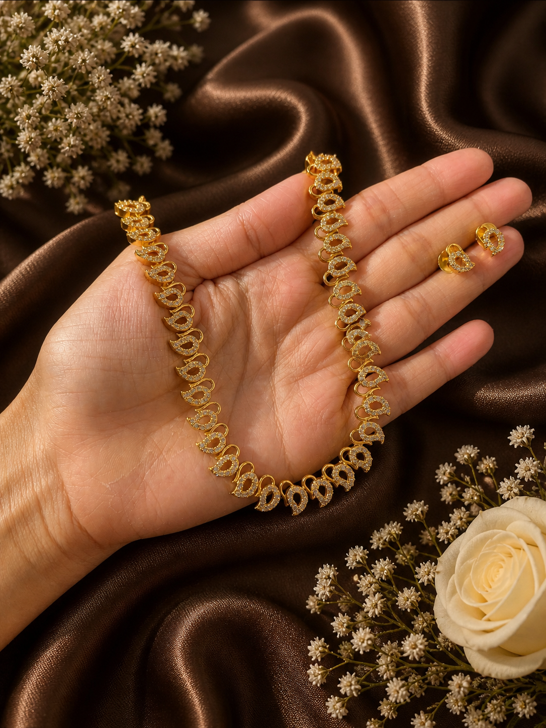 A hand cradling a gold-plated necklace and small matching stud earrings, designed with a continuous string of traditional paisley or mango motifs embedded with micro-pave white CZ stones on a brown satin background.