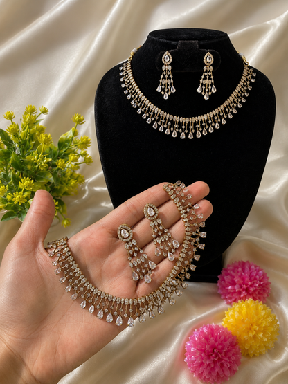 A matching gold-plated jewelry set displayed on a hand and a black bust, featuring a collar necklace with a fringe of vertical bars and dangling teardrop-cut white CZ stones, paired with multi-tier teardrop drop earrings.