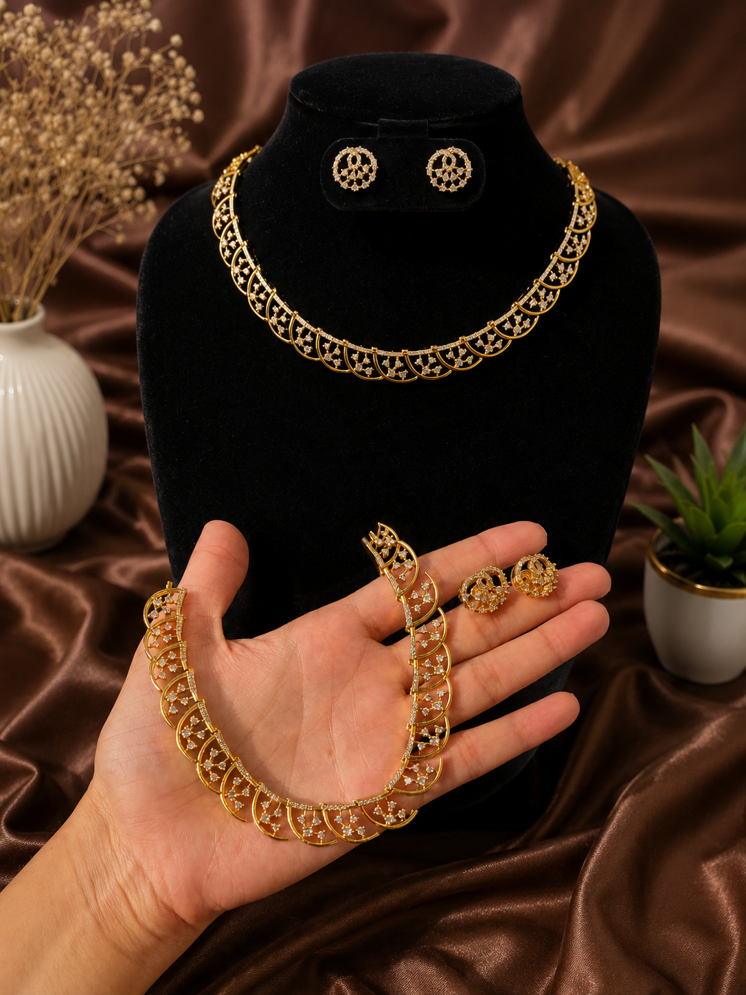 Elegant gold-plated jewelry set featuring a collar necklace with a delicate scalloped, geometric openwork design studded with clear CZ crystals. One set is displayed on a black velvet bust with matching circular stud earrings, while another identical set rests in a hand against a brown satin backdrop.