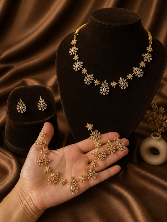 Gold-plated choker jewelry set featuring starburst floral motifs set with clear cubic zirconia crystals. One necklace is displayed on a brown velvet bust, matching stud earrings rest on a smaller stand, and a duplicate set is held in a person's hand against a brown satin background.