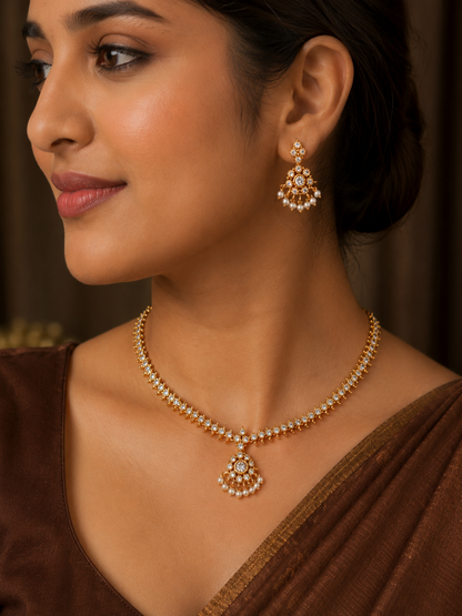 Close-up of a woman in a brown saree wearing a delicate gold-plated choker necklace studded with small clear crystals, featuring a central cluster pendant with hanging mini pearls and matching drop earrings.