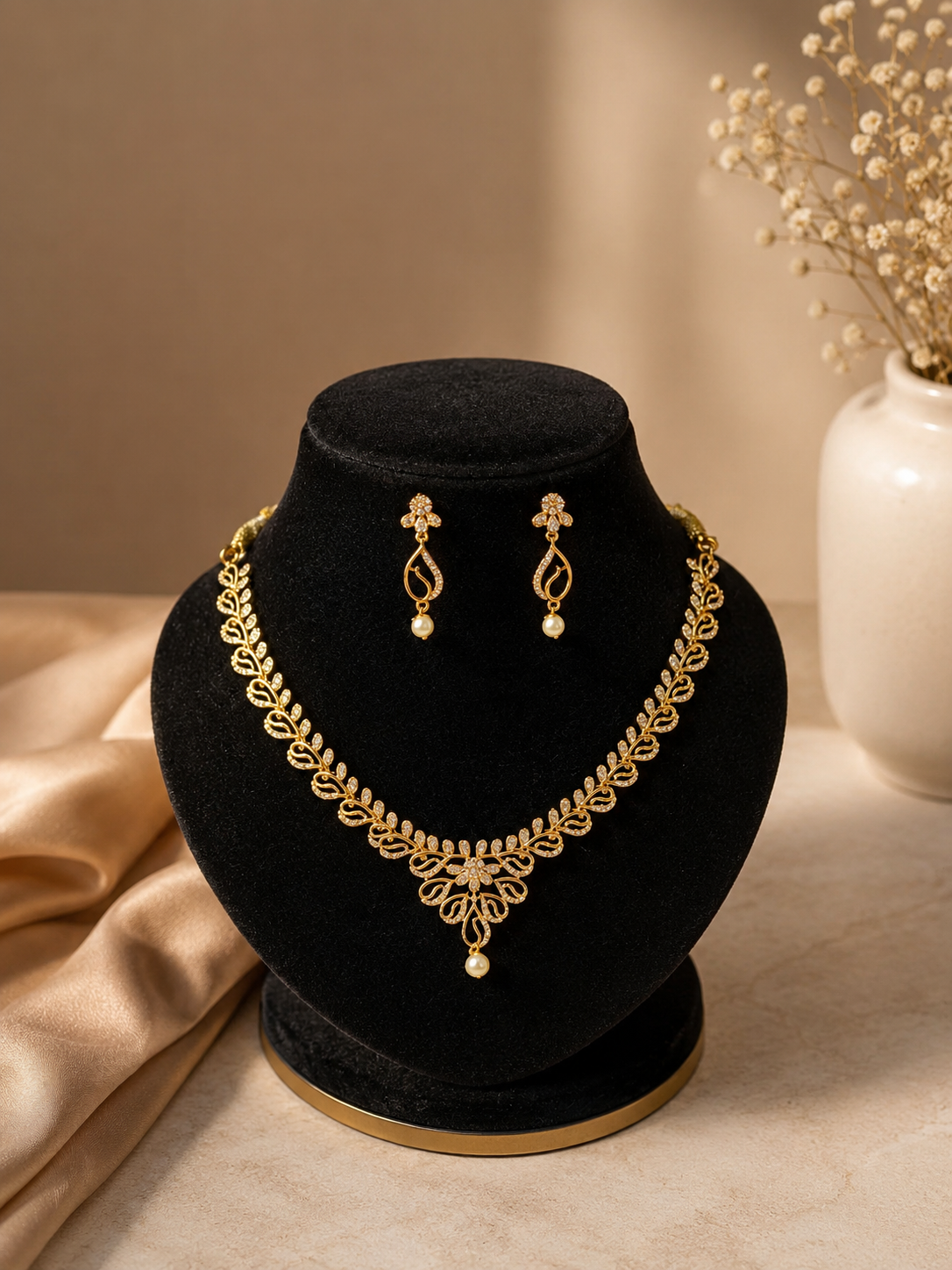 Elegant gold-plated jewelry set featuring a necklace with a continuous vine and leaf motif embedded with clear CZ crystals, meeting at a central decorative drop with a single hanging pearl. The set is displayed on a black velvet bust with matching drop earrings against a warm beige background.