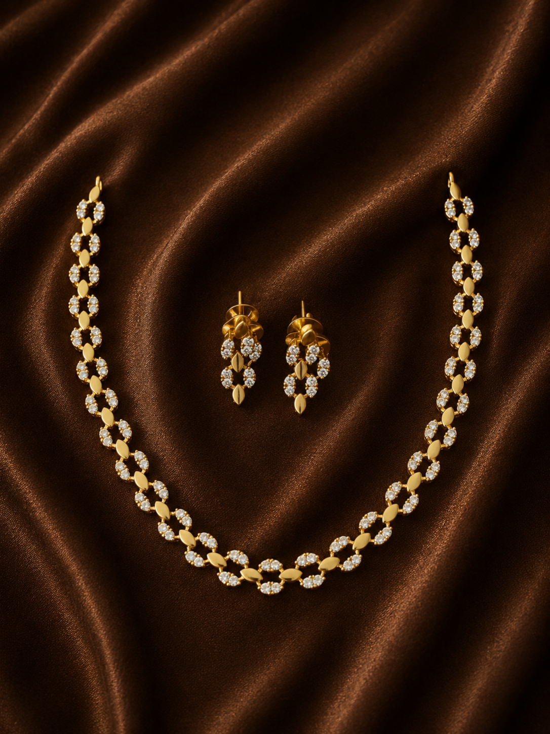 A luxury gold necklace and matching drop earrings set adorned with shimmering white crystals, elegantly arranged on a draped brown satin fabric background.