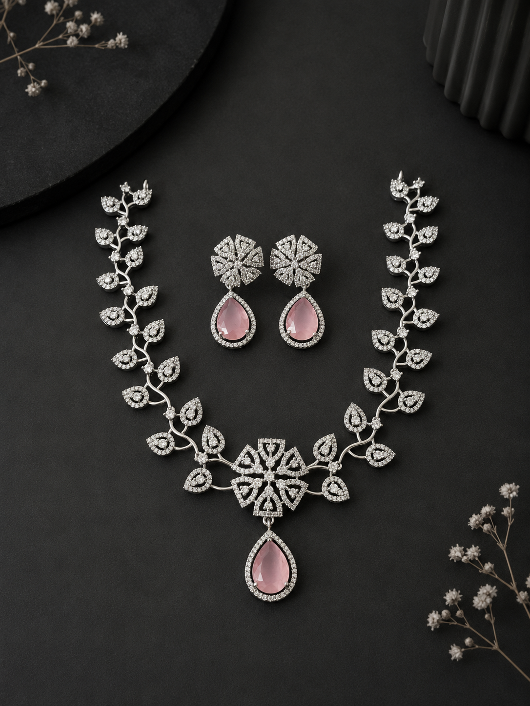 A silver-toned floral necklace and matching earrings set featuring crystal-studded leaves and prominent pink teardrop gemstones, displayed on a sleek black background.