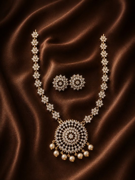 A gold-plated necklace with a prominent, round crystal-encrusted medallion pendant adorned with dangling pearls, paired with matching round stud earrings on a draped brown satin fabric.