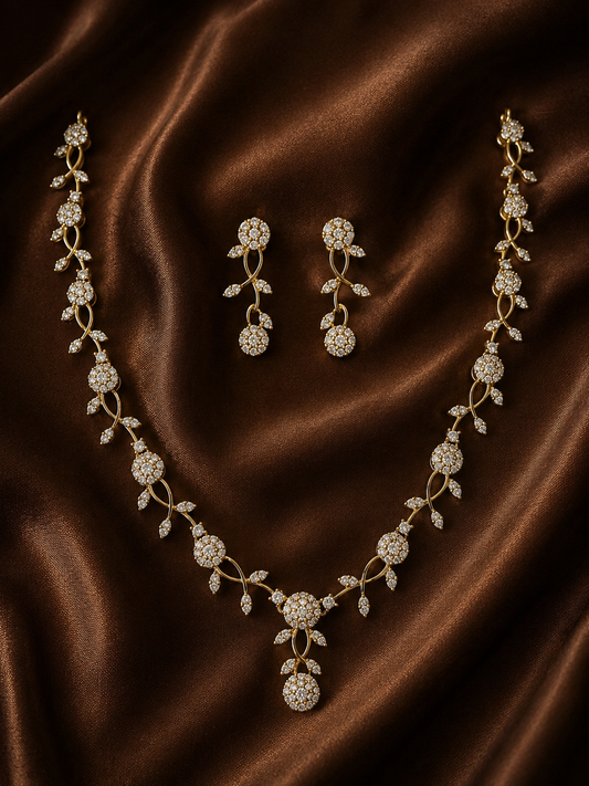 A gold-plated necklace and matching drop earrings set with a delicate interwoven vine design and round, crystal-studded floral motifs, resting on a draped brown satin fabric.
