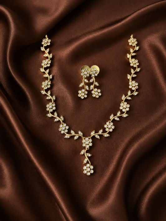 A gold-plated necklace and matching earrings set featuring an intricate flower and leaf vine design with embedded crystal stones, displayed on a wavy brown satin fabric.