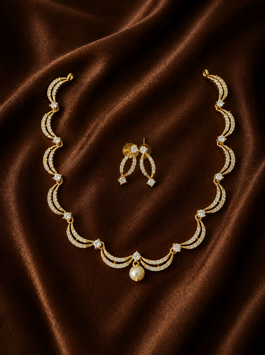 A gold-plated necklace and matching curved drop earrings set, featuring brilliant crystal-encrusted crescent shapes and a single hanging pearl pendant, arranged on a draped brown satin fabric.