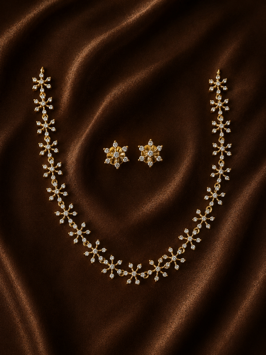 A gold-plated necklace chain and matching stud earrings set, featuring brilliant crystal-studded starburst and flower designs, arranged on a rich, draped brown satin fabric background.