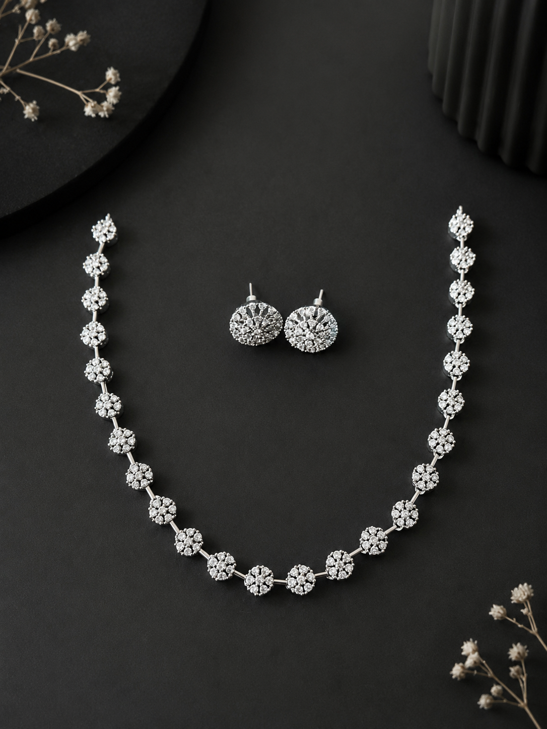 A silver-plated necklace and matching round stud earrings set featuring delicate floral motifs encrusted with sparkling white crystals, showcased on a minimalist matte black background.