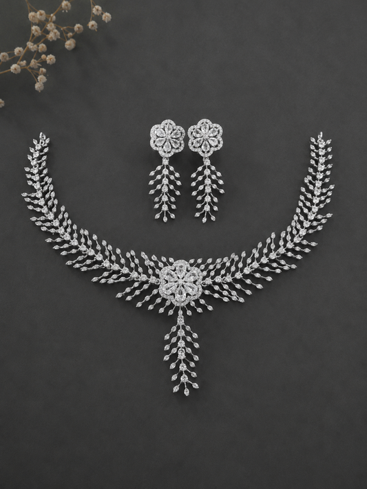 A premium silver-toned cubic zirconia jewelry set featuring a detailed floral centerpiece necklace with a hanging leaf tendril and matching dangle earrings on a dark grey background.