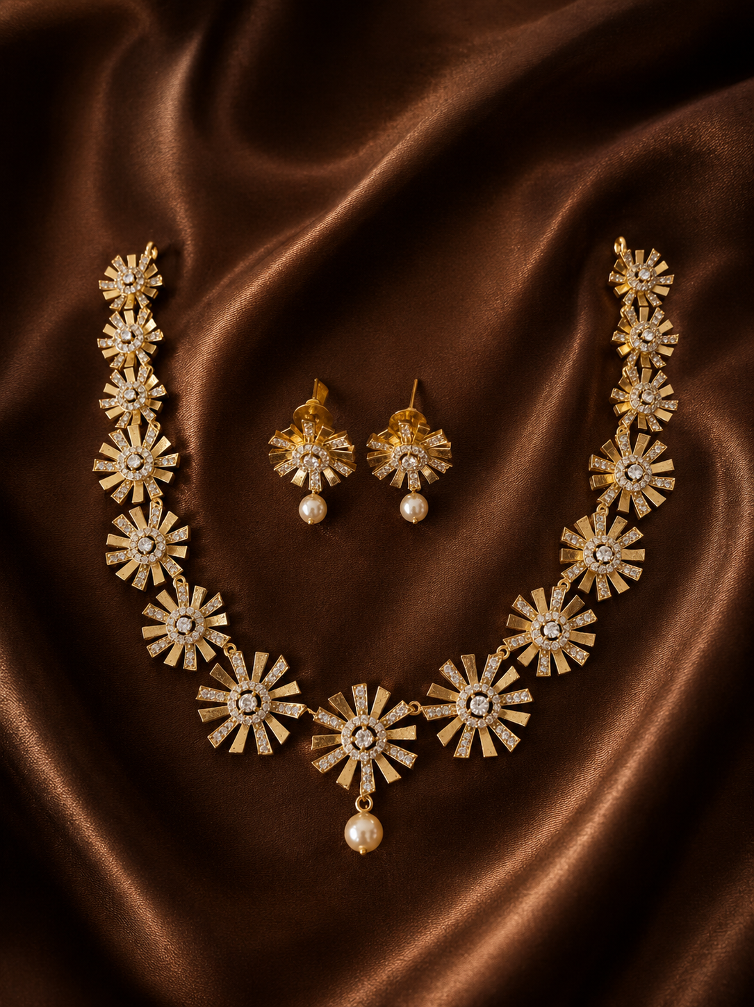 A gold-plated choker necklace and matching stud earrings featuring a textured sunburst or starburst motif with crystal centers and teardrop pearls, arranged on brown satin fabric.