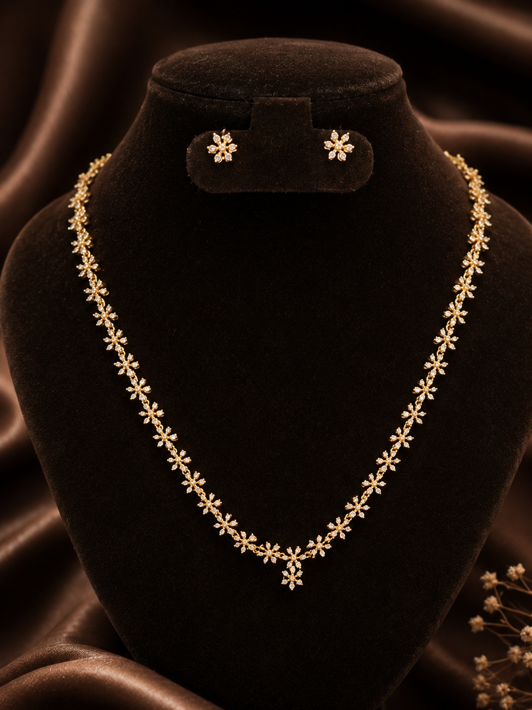 A dainty gold-plated necklace chain and matching small flower stud earrings embellished with fine crystals, displayed on a black velvet bust mannequin against brown silk.