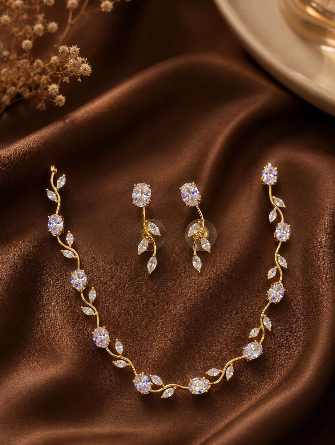 Vanitha minimalist jewelry set featuring a delicate wavy gold-toned chain necklace adorned with alternating oval crystals and marquise-cut crystal leaf accents, displayed flat on rich brown satin fabric alongside matching dangling leaf-style drop earrings.