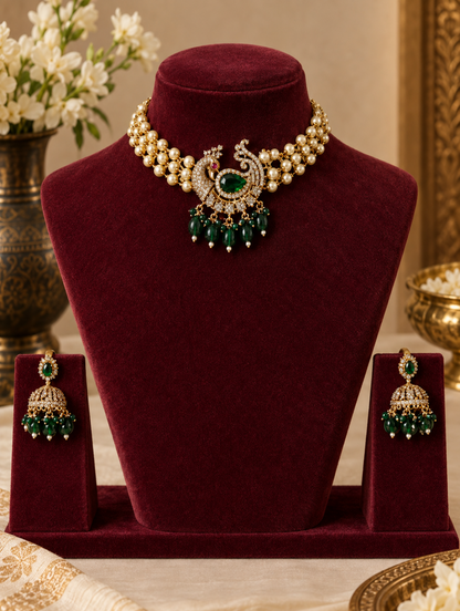A traditional Indian jewelry set displayed on a deep burgundy velvet jewelry stand. The set includes a pearl and gold-plated choker necklace with a central peacock motif featuring a green stone and dangling green beads, along with matching green and gold jhumka earrings.