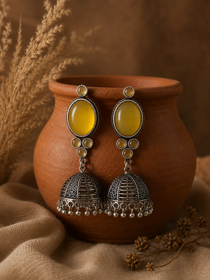 Intricately designed oxidized silver Jhumka earrings featuring large, smooth yellow oval stones, displayed hanging on an earthen clay pot in the file E002_Sitara_299.png.