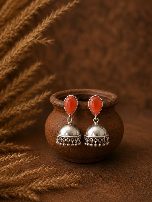 A pair of polished silver Jhumka earrings featuring vibrant orange teardrop stones, hanging on a small brown terracotta pot as seen 