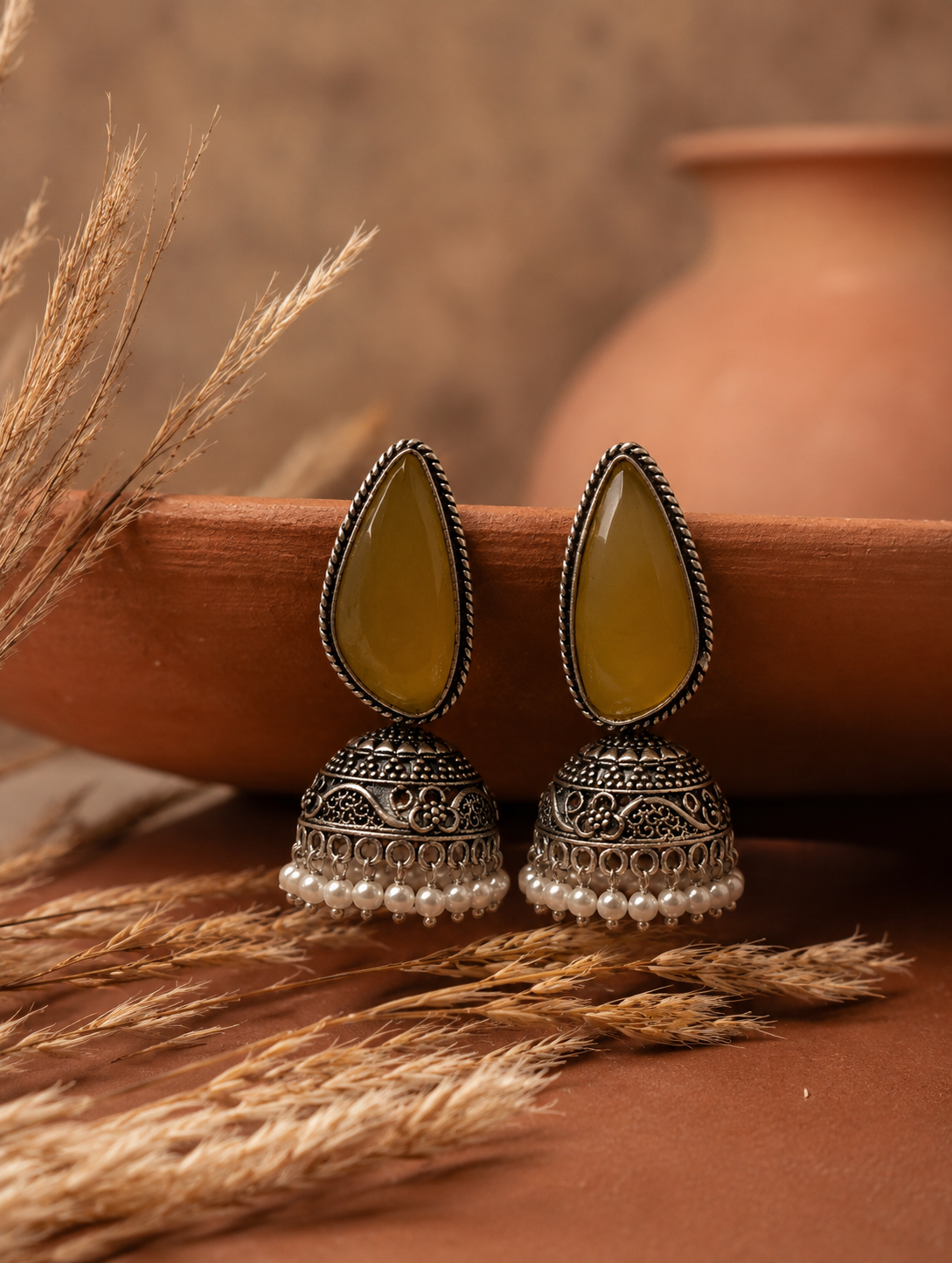 A pair of oxidized silver Morni Jhumka earrings featuring large olive-yellow teardrop stones and a fringe of white pearl beads, resting against a terracotta plate as shown