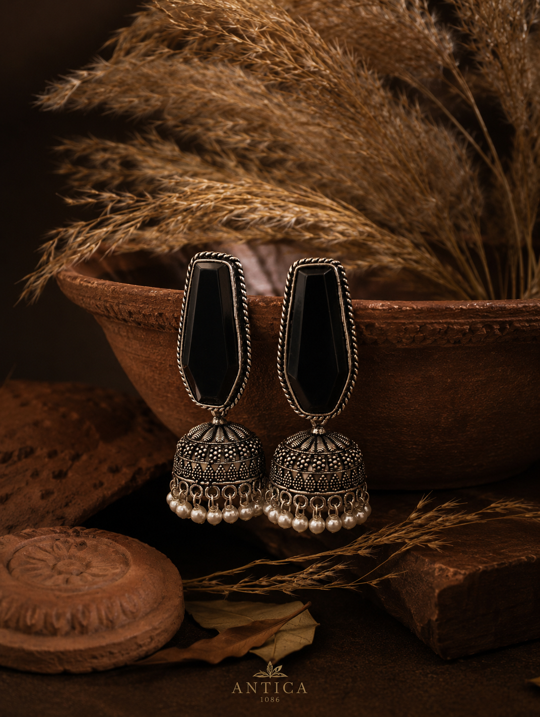 A pair of oxidized silver Noorani Jhumka earrings featuring large, elongated geometric black stones and dangling white pearl beads, hanging on the rim of a terracotta bowl as shown
