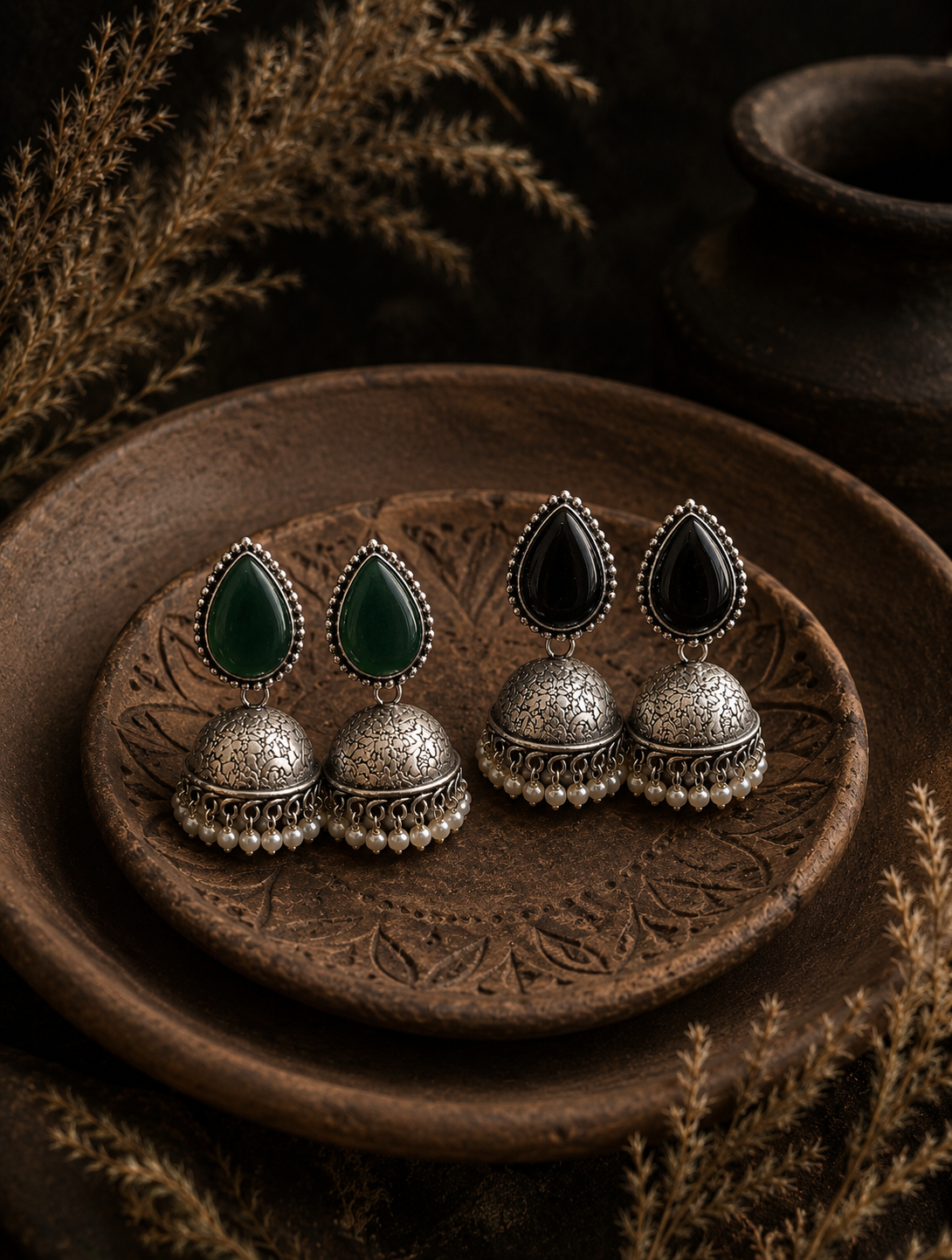 Two pairs of oxidized silver Roohi Jhumka earrings, one with dark green teardrop stones and the other with black teardrop stones, decorated with small white pearls and resting on a carved terracotta plate