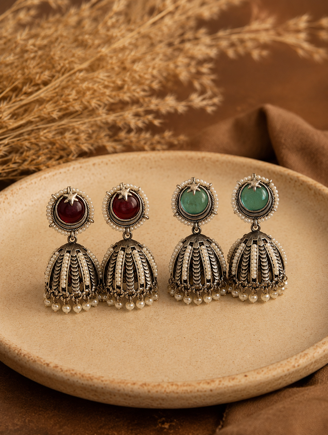 Two pairs of oxidized silver Kohinoor Jhumka earrings, one with round ruby red stones and the other with mint green stones, featuring star motifs and elaborate vertical pearl detailing on the domes, displayed on a beige plate 