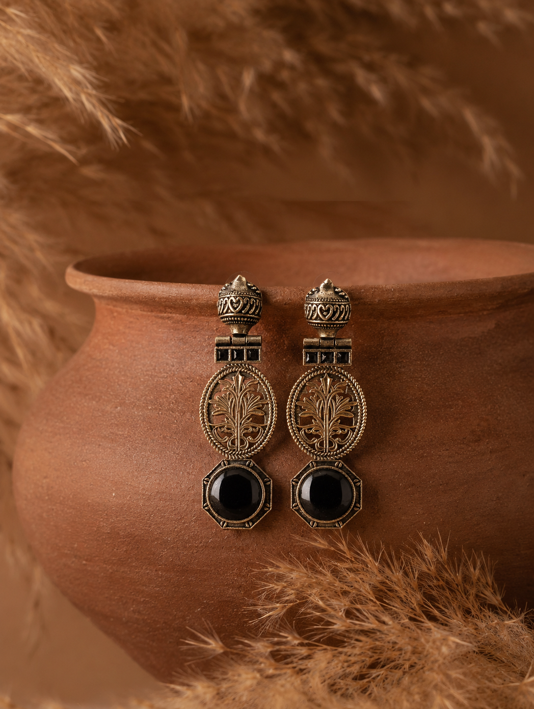 A pair of antique oxidized Kirana drop earrings featuring detailed circular tree motifs and bottom octagonal settings with smooth black stones, shown hanging on a rustic terracotta pot.