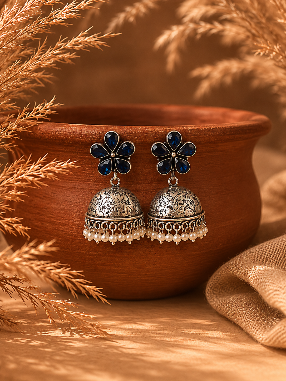 A pair of oxidized silver Pallu Jhumka earrings featuring dark blue gemstone flower studs, finely engraved floral domes, and dangling white pearl beads, displayed hanging on a terracotta pot 