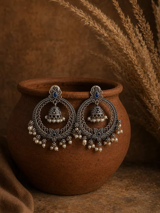 A pair of large, circular oxidized silver Lallu hoop earrings featuring an internal hanging jhumka dome, floral details with blue stone accents, and small white dangling pearls, draped over a brown clay pot