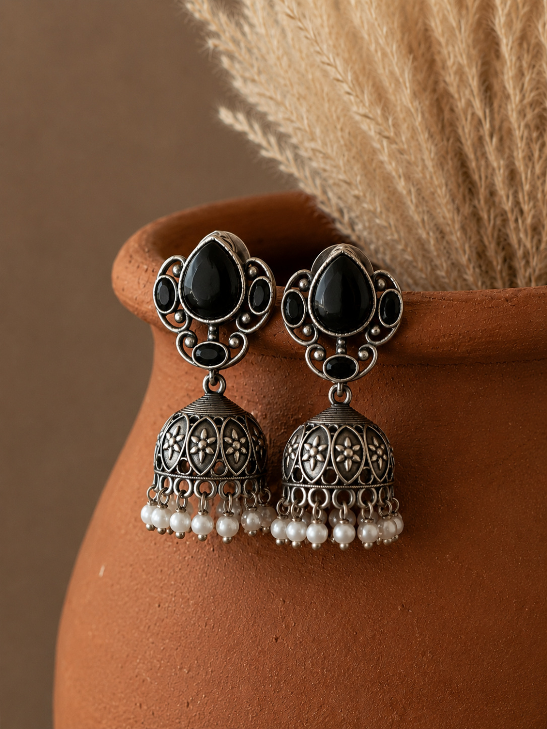 A pair of oxidized silver Poonam Jhumka earrings featuring black teardrop stones in a decorative scrollwork stud, detailed floral-embossed domes, and a fringe of dangling white pearl beads, displayed hanging on a terracotta pot