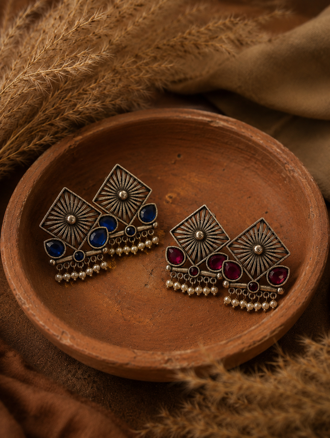 Pair of Mina oxidized silver tribal earrings with geometric starburst designs, blue and ruby red stone accents, and small hanging pearls, displayed in a rustic clay bowl.