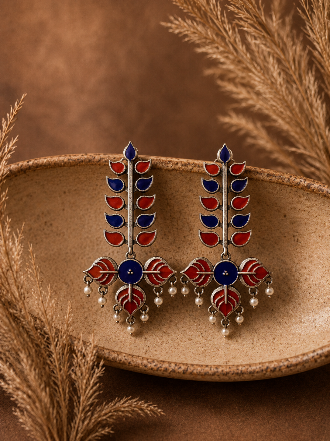 Decorative earrings with red, blue, and silver design on a textured surface with dried plants.