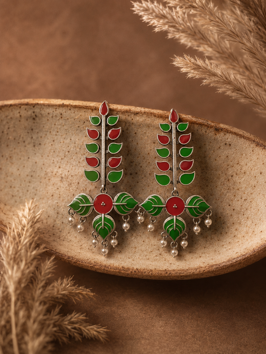 A pair of Lilly oxidized silver earrings with a vertical tree or leaf motif filled with red and green enamel, dangling small white pearls, displayed on a rustic ceramic dish.