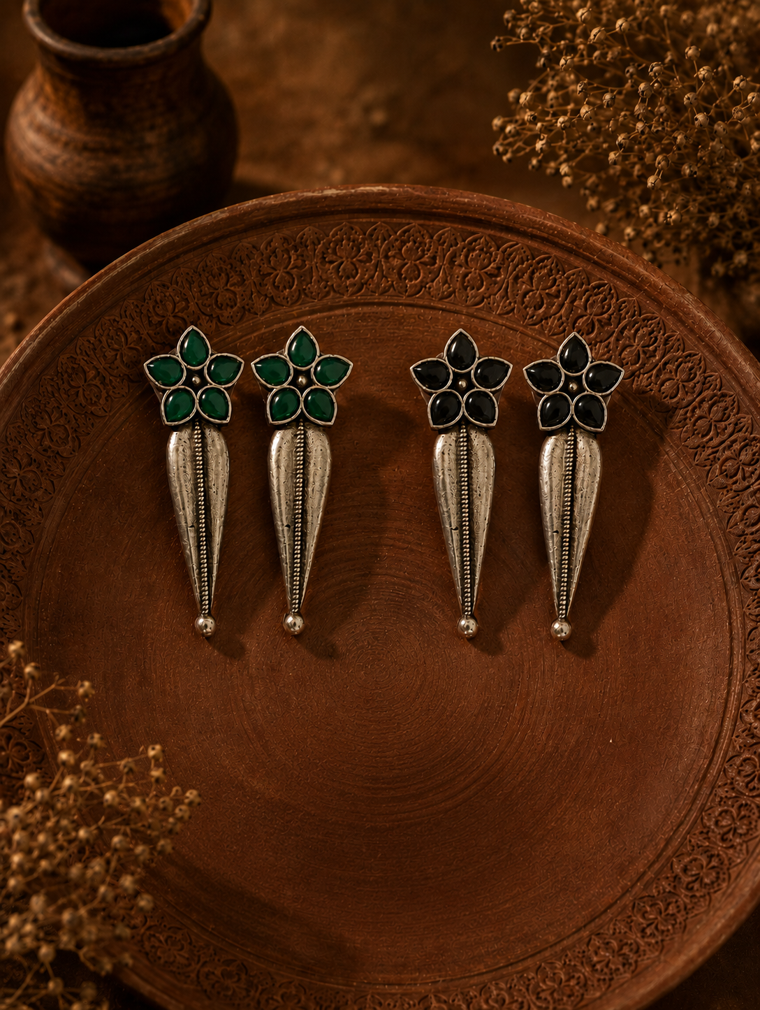 Two pairs of Swara oxidized silver dangle earrings, one with emerald green flower studs and the other with black flower studs, both featuring sleek elongated metal drops on a carved clay plate.