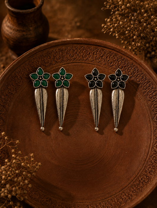 Two pairs of Swara oxidized silver dangle earrings, one with emerald green flower studs and the other with black flower studs, both featuring sleek elongated metal drops on a carved clay plate.