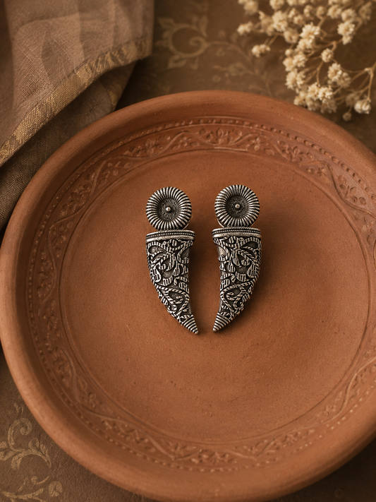 A pair of Srinika oxidized silver tribal earrings featuring a textured, spiral circular top and an engraved, curved horn-shaped drop with detailed filigree patterns, resting in a clay plate.