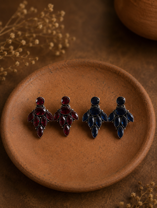 Two pairs of Uma oxidized silver cluster earrings, one set featuring ruby red navette gemstones and the other featuring deep sapphire blue gemstones, displayed on a small clay dish.