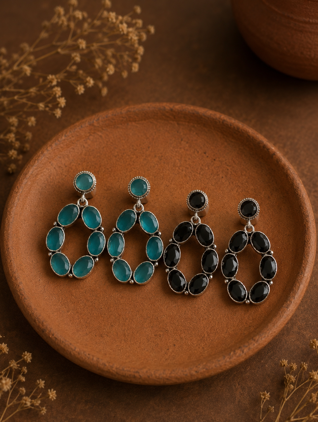 Two pairs of Karishma silver open-oval hoop earrings, one set featuring translucent sea green stones and the other featuring glossy black stones, arranged on a clay dish.
