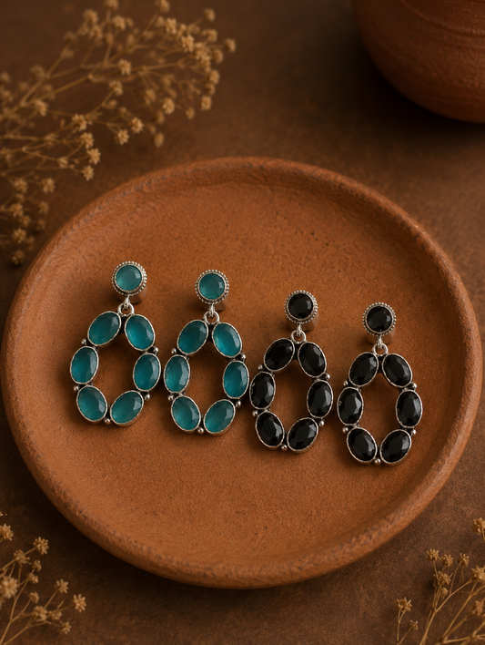 Two pairs of Karishma silver open-oval hoop earrings, one set featuring translucent sea green stones and the other featuring glossy black stones, arranged on a clay dish.