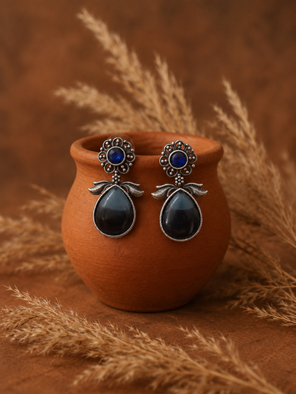 A pair of Pramila oxidized silver drop earrings featuring a circular floral filigree stud with a round sapphire blue stone center and a large, smooth black teardrop dangling from a terracotta pot.