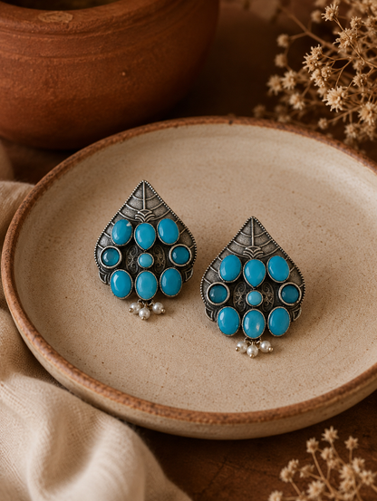 A pair of Chitra oxidized silver oversized shield-shaped stud earrings featuring smooth, oval turquoise stone clusters and small hanging pearl droplets, displayed on a ceramic dish.