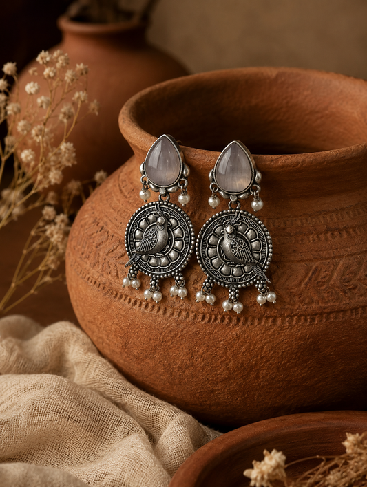 A pair of Savi oxidized silver drop earrings featuring translucent pink teardrop stone studs, a circular textured medallion with a bird or parrot motif, and small hanging pearl clusters, resting on a clay pot.