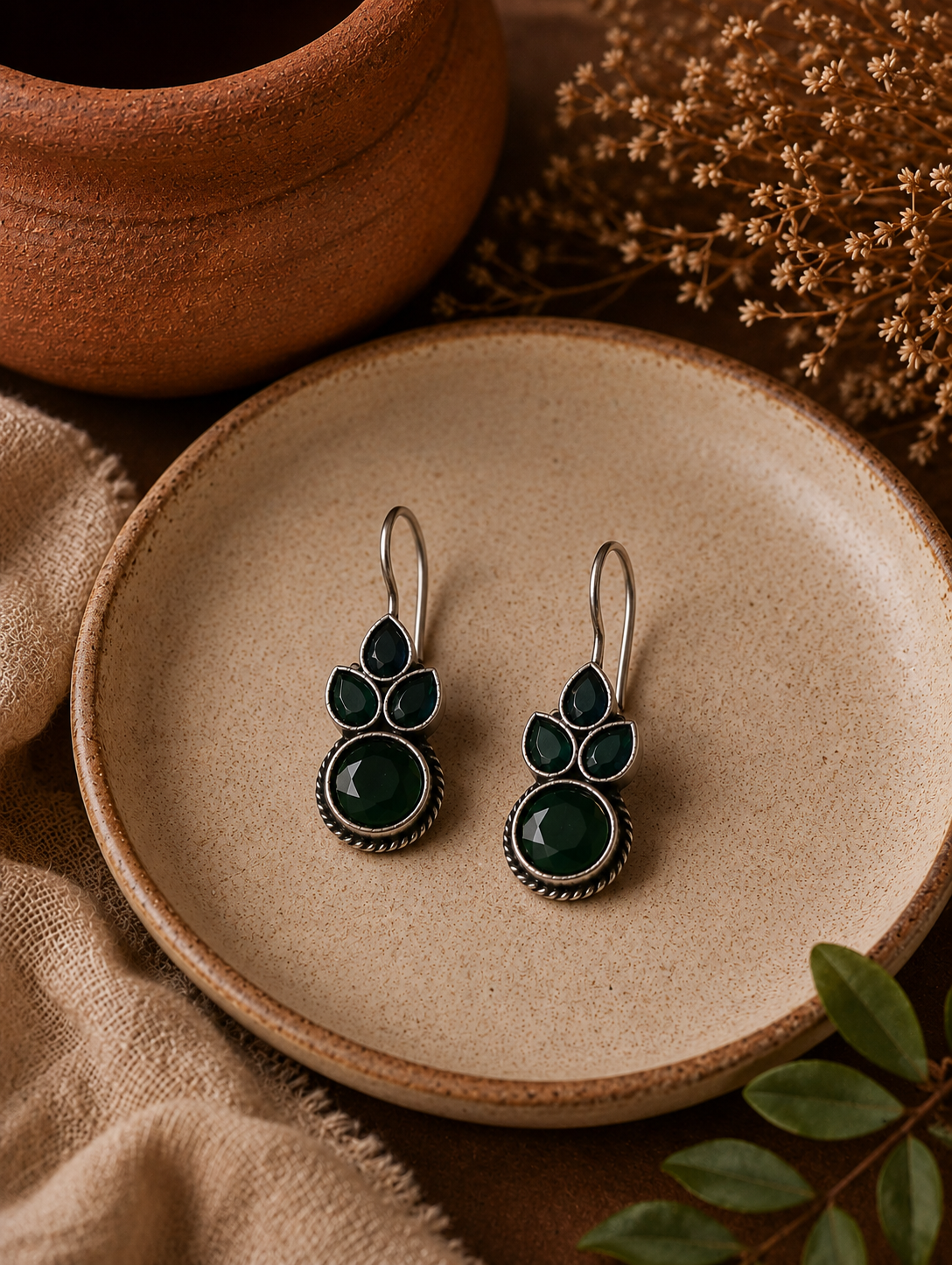 A pair of Seema silver drop earrings with elegant hook closures, featuring a cluster of dark green teardrop stones sitting above a large, round faceted emerald green stone, displayed on a ceramic plate.