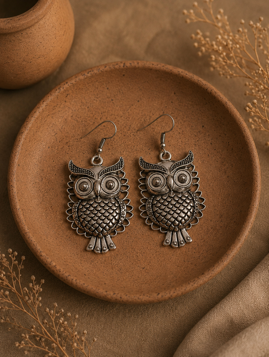 A pair of Parijatha oxidized silver dangle earrings featuring a detailed owl design with big round eyes, textured scales on the body, and pointed horns, placed inside a terracotta dish.