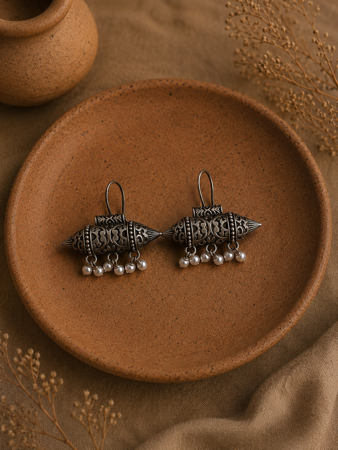 A pair of Sashi oxidized silver tribal earrings featuring an elongated, barrel or capsule shape with detailed ornate cutouts and tiny hanging white pearls, displayed on a clay plate.