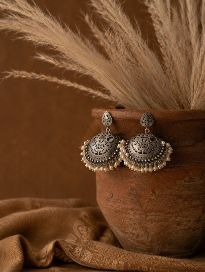 A pair of large, dome-shaped oxidized silver statement earrings featuring intricate openwork filigree carvings and a thick fringe of hanging seed pearls, displayed on a ceramic plate.